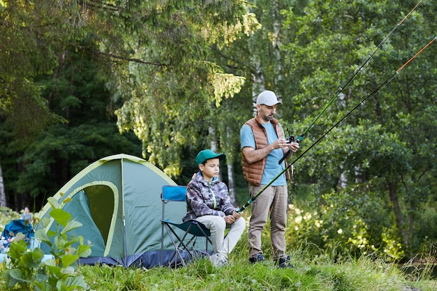 Retrato de cuerpo entero de amoroso padre e hijo pescando juntos en el lago durante el viaje de campamento en la naturaleza, espacio de copia