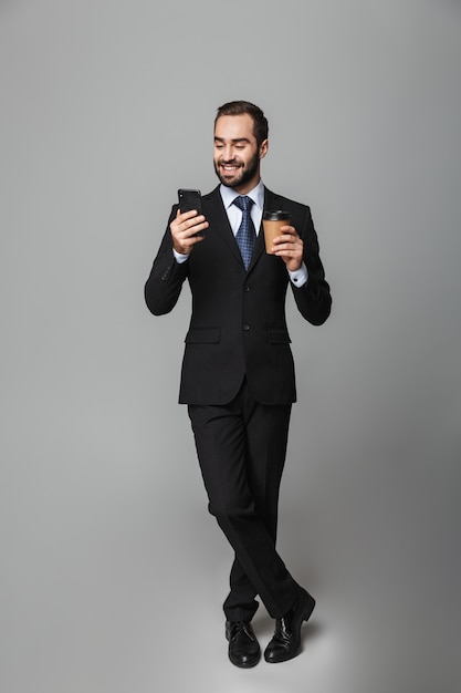 Retrato de cuerpo entero de un apuesto hombre de negocios confiado con traje aislado, sosteniendo café para llevar, usando teléfono móvil