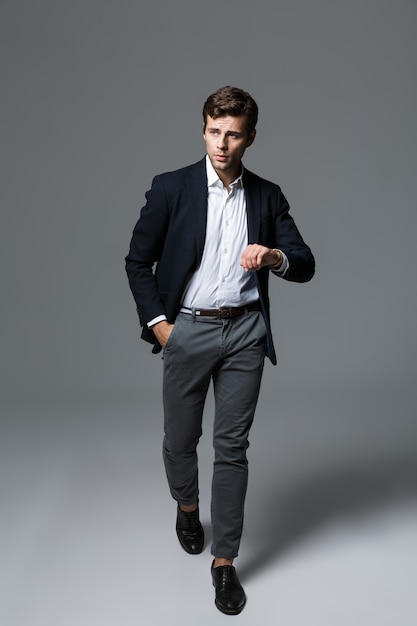 Retrato de cuerpo entero de un apuesto joven empresario vestido con traje aislado sobre pared gris, posando, comprobando el tiempo, caminando