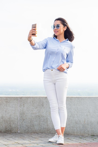 Retrato de cuerpo entero de una atractiva mujer morena haciendo selfie en la calle al aire libre del teléfono inteligente