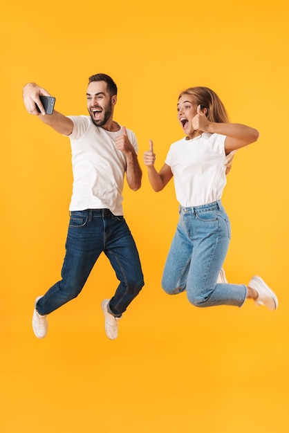 Foto retrato de cuerpo entero de una atractiva pareja joven alegre vistiendo ropa casual tomando un selfie mientras salta aislado sobre pared amarilla