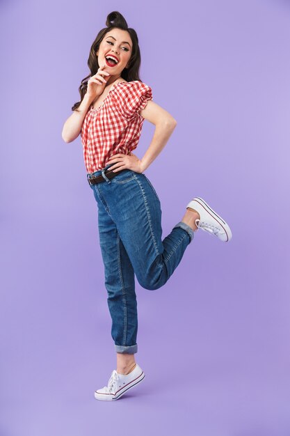 Retrato de cuerpo entero de una chica pin-up coqueteando en ropa de estilo americano sonriendo a la cámara aislada sobre la pared violeta