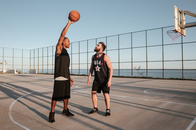 Retrato de cuerpo entero de dos hombres jóvenes jugando baloncesto en el patio de recreo