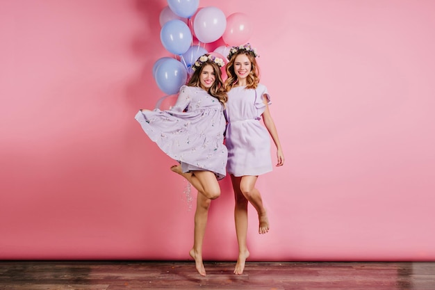 Retrato de cuerpo entero de dos mujeres descalzas que se divierten en la fiesta. Foto interior de chicas caucásicas en vestidos bailando con globos.