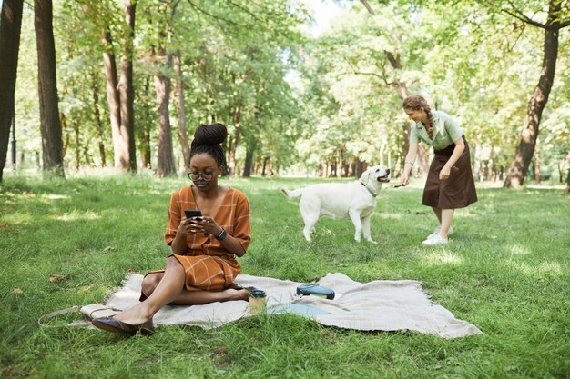 Retrato de cuerpo entero de dos mujeres jóvenes en el parque jugando con el perro al aire libre y usando el teléfono inteligente, espacio de copia