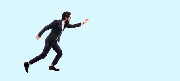 Retrato de cuerpo entero del empresario barbudo y moderno que se ejecuta contra la pared azul, el hombre tira de la mano hacia adelante para atrapar algo, imagen panorámica