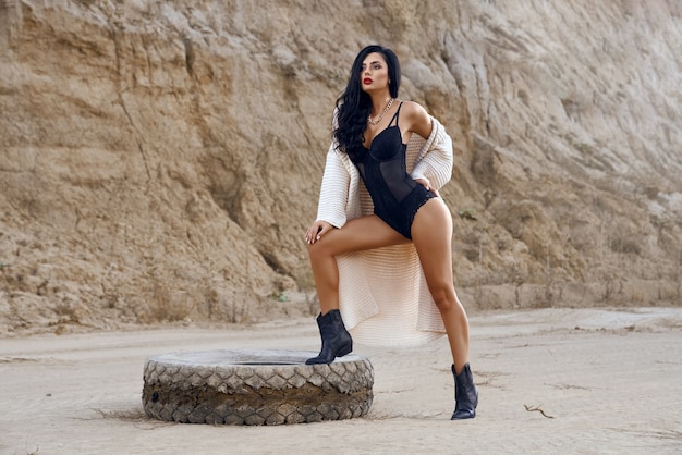 Retrato de cuerpo entero de una encantadora modelo femenina con cabello largo y oscuro vestida con ropa de cuerpo de moda, posando en el desierto cerca de la llanta vieja