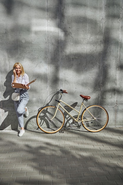 Retrato de cuerpo entero de una encantadora mujer rubia caucásica sosteniendo una carpeta de papel con documentos en la mano mientras llama a sus clientes