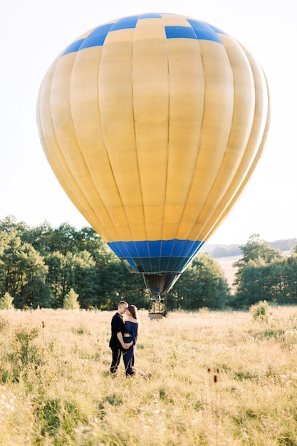 Retrato de cuerpo entero de una encantadora pareja joven vestida de negro, abrazándose y disfrutando de la caminata de verano en el campo, esperando su recorrido en globo aerostático