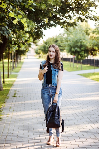 Foto retrato de cuerpo entero de una estudiante con chaleco de mezclilla y jeans pasando tiempo al aire libre en verano