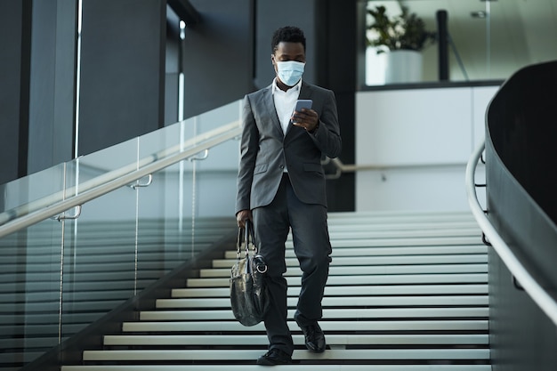 Retrato de cuerpo entero del exitoso empresario afroamericano con máscara y usando el teléfono inteligente mientras camina hacia la cámara en el edificio de oficinas, espacio de copia