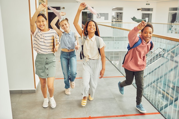 Retrato de cuerpo entero de un grupo multiétnico de escolares sosteniendo máscaras y sonriendo felizmente mientras camina hacia la cámara en el interior