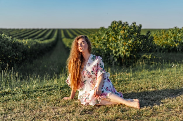 Retrato de cuerpo entero de hermosa mujer con pelo largo y rizado en campo de grosella. mujer en un vestido ligero se sienta en la hierba en un día soleado