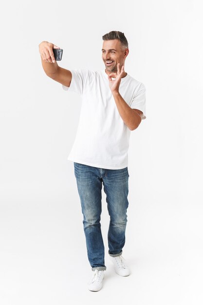 Retrato de cuerpo entero de un hombre sin afeitar de 30 años con camiseta casual y jeans tomando una foto selfie en el teléfono móvil mientras sostiene en la mano aislado en blanco