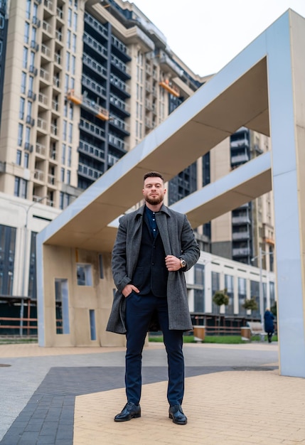 Retrato de cuerpo entero de un hombre de negocios feliz de pie al aire libre. Abrigo y traje elegante. Fondo de edificio moderno.