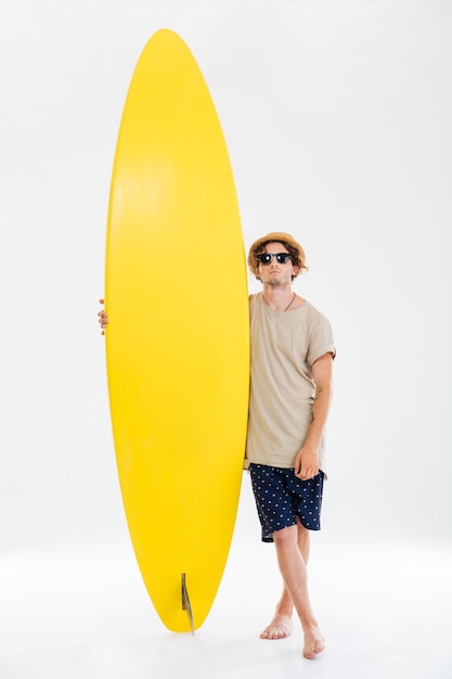 Retrato de cuerpo entero de un hombre de playa con gafas de sol y sombrero con tabla de surf aislada en la pared blanca