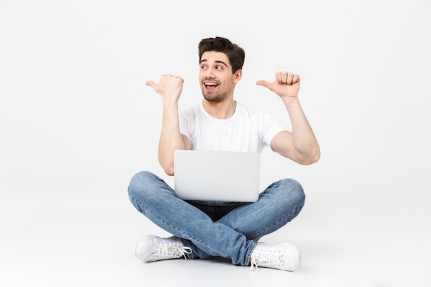 Retrato de cuerpo entero de un joven alegre con ropa casual aislado sobre blanco, sentado con una computadora portátil, apuntando hacia afuera