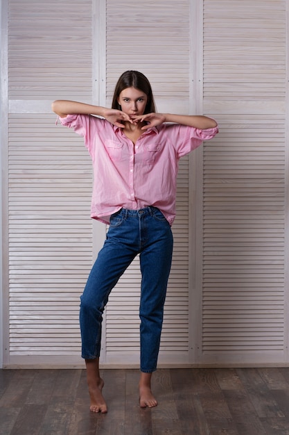 Retrato de cuerpo entero de la joven modelo de moda viste jeans azules y camiseta rosa posando en el fondo de las contraventanas