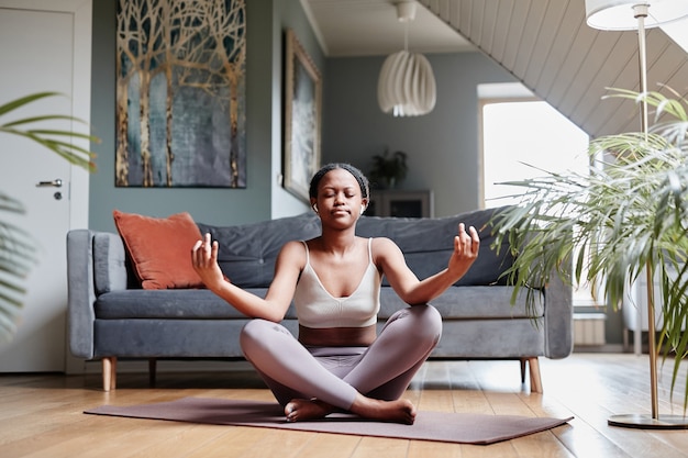 Retrato de cuerpo entero de joven mujer afroamericana meditando en casa mientras está sentado en posición de loto ...