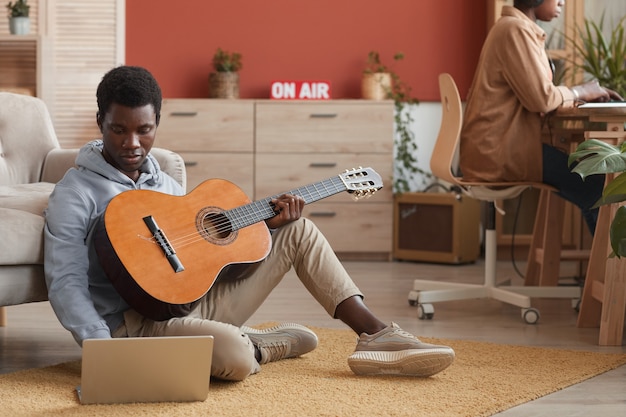 Retrato de cuerpo entero de un joven músico afroamericano tocando la guitarra y usando la computadora portátil mientras está sentado en el piso en el estudio de grabación, espacio de copia