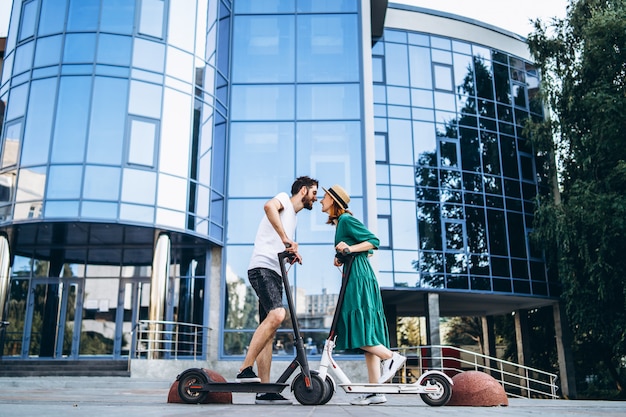 Retrato de cuerpo entero de una joven pareja romántica con scooters eléctricos, caminando en la ciudad. Mujer joven con sombrero y hombre disfruta de un paseo