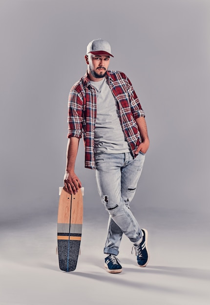 Retrato de cuerpo entero de un joven patinador apoyado en su patineta y mirando a la cámara aislada sobre fondo gris.