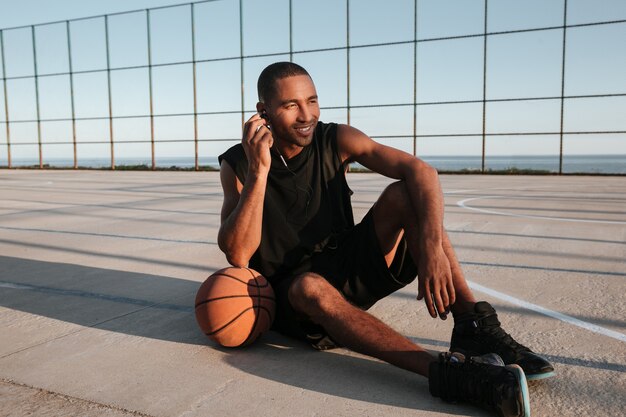 Retrato de cuerpo entero de un jugador de baloncesto sonriente sentado en el patio de recreo y escuchando música con auriculares