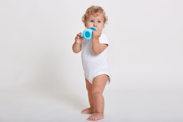 Retrato de cuerpo entero de lindo niño bebiendo agua de botella, niño de un año jugando con taza de bebé, adorable niño de pelo rizado mirando a otro lado