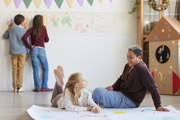 Retrato de cuerpo entero de una maestra sonriente sentada en el piso con niños haciendo dibujos mientras disfruta de la clase de arte en Navidad, espacio de copia