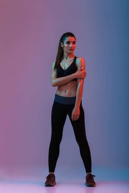 Retrato de cuerpo entero de mujer atlética en ropa deportiva posando durante ejercicios aeróbicos, aislado sobre la pared púrpura