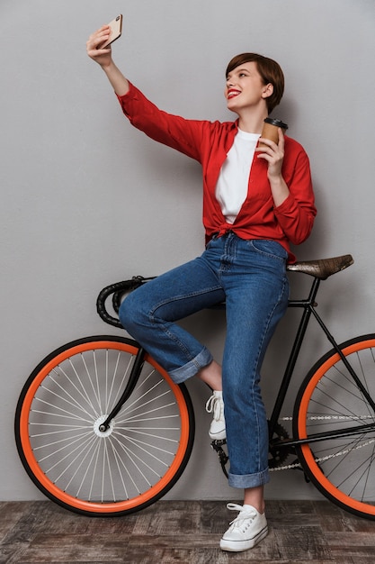 Retrato de cuerpo entero de mujer complacida tomando selfie foto en smartphone de pie en bicicleta con taza de café aislada sobre pared gris