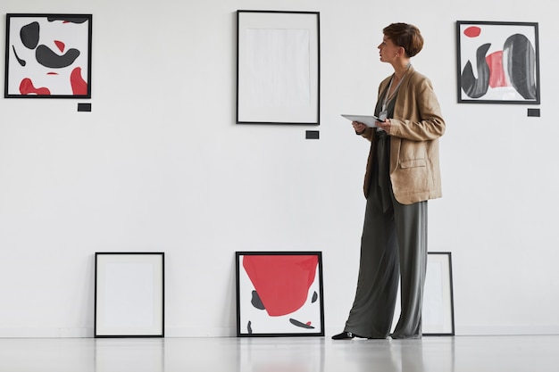 Retrato de cuerpo entero de una mujer elegante creativa mirando pinturas colgadas en la pared blanca mientras visita la exposición de la galería de arte moderno,