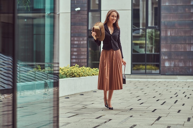 Retrato de cuerpo entero de una mujer elegante de moda feliz que lleva una chaqueta negra y una falda sostiene un sombrero y un embrague de bolso caminando en un centro de la ciudad europea.