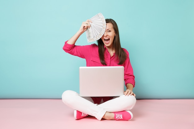 Retrato de cuerpo entero de una mujer joven en blusa de camisa rosa, pantalón blanco sentado en el piso con dinero pc