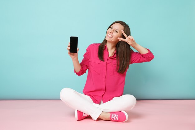 Retrato de cuerpo entero de una mujer joven divertida en camisa rosa, pantalones blancos sentarse en el piso, sujetar el teléfono celular