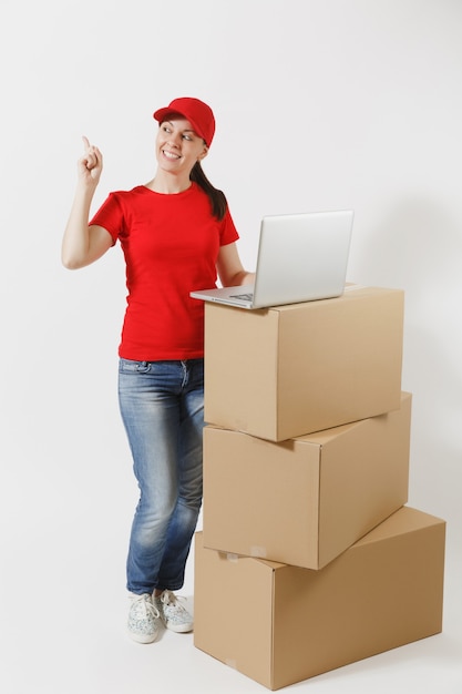 Retrato de cuerpo entero de mujer joven de entrega en gorra roja, camiseta aislada sobre fondo blanco. Mensajero mujer de pie cerca de cajas de cartón vacías con ordenador portátil. Recibiendo paquete. Copia espacio