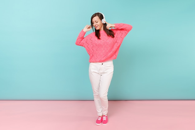 Retrato de cuerpo entero de mujer joven feliz en suéter rosa tejido, pantalón blanco, auriculares