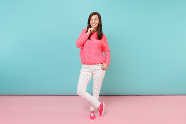 Retrato de cuerpo entero de mujer joven sonriente en suéter rosa de punto, pantalones blancos posando