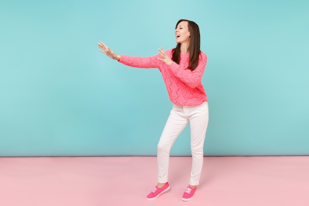 Retrato de cuerpo entero de mujer joven sonriente en suéter rosa de punto, pantalones blancos posando