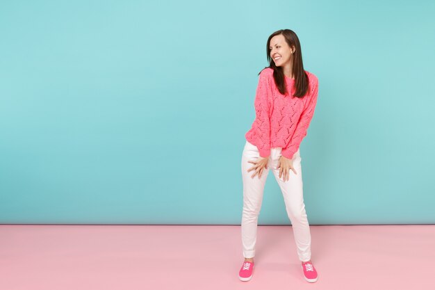 Retrato de cuerpo entero de mujer joven sonriente en suéter rosa de punto, pantalones blancos posando