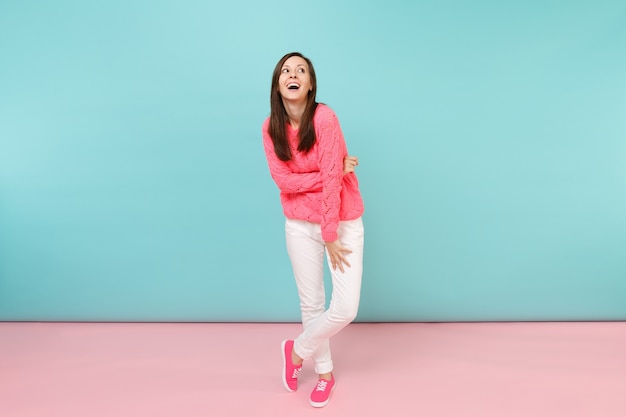 Retrato de cuerpo entero de mujer joven sonriente en suéter rosa de punto, pantalones blancos posando