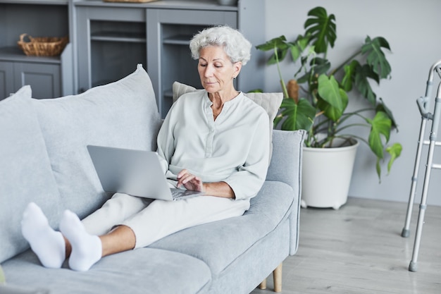 Retrato de cuerpo entero de una mujer mayor moderna que usa la computadora portátil mientras está acostado en el sofá en el espacio de copia de casa