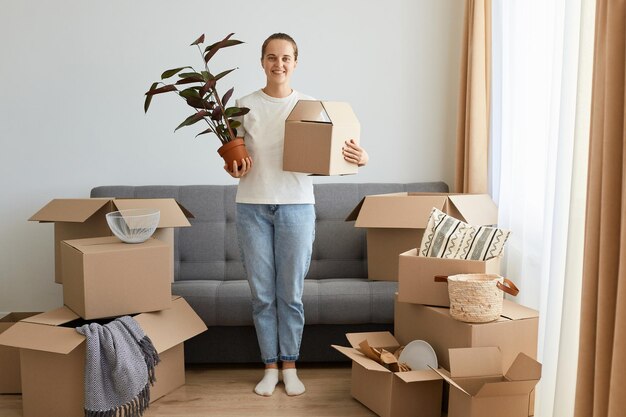 Retrato de cuerpo entero de una mujer positiva sonriente con camiseta blanca y jeans de pie en su nuevo hogar sosteniendo una maceta con una planta y una caja de cartón