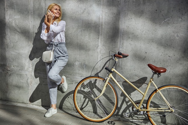 Retrato de cuerpo entero de mujer riendo de pie cerca de su bicicleta mientras conversa con un socio comercial en el teléfono móvil