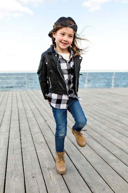 Retrato de cuerpo entero de una niña encantadora vestida con gorra y chaqueta de cuero divirtiéndose y posando cerca del mar