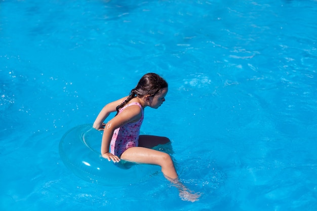 Niña linda en traje de baño azul sosteniendo una tabla de natación de  plástico rosa mientras está de pie frente a la piscina y pone los dedos de  los pies en el