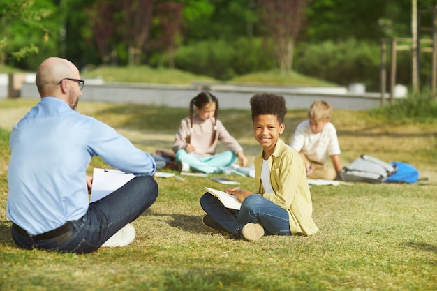 Retrato de cuerpo entero de un niño afroamericano sonriente sentado en la hierba verde y mirando a la cámara mientras disfruta de la clase al aire libre en la luz del sol, espacio de copia