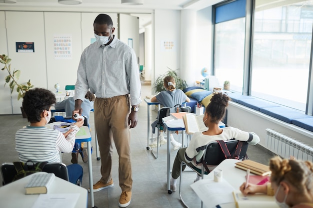 Retrato de cuerpo entero del profesor afroamericano desinfectando las manos de los niños en el aula de la escuela, espacio de copia