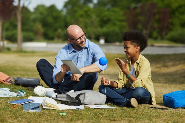 Retrato de cuerpo entero del profesor calvo sonriente hablando con un niño afroamericano que sostiene el planeta modelo mientras disfruta de una lección de astronomía al aire libre en la luz del sol, espacio de copia