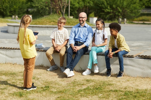 Retrato de cuerpo entero del profesor sonriente escuchando a una niña dando una presentación mientras está sentado con un grupo de niños y disfruta de una lección al aire libre en la luz del sol